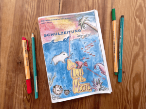 Auf einem Holztisch liegt eine gedruckte Ausgabe der Schulzeitung mit dem Titel „Die Welt der Meere“, sowie vier Stifte