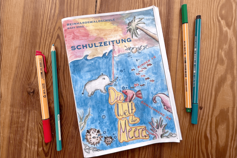 Auf einem Holztisch liegt eine gedruckte Ausgabe der Schulzeitung mit dem Titel „Die Welt der Meere“, sowie vier Stifte