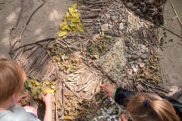 Projekt_LandArt_2023_2 Das Bild zeigt einen Blick auf sandigen Boden. Von oben sind zwei Kinder zu sehen, die ein Muster aus Pflanzenteilen, Stöcken und Blättern legen.