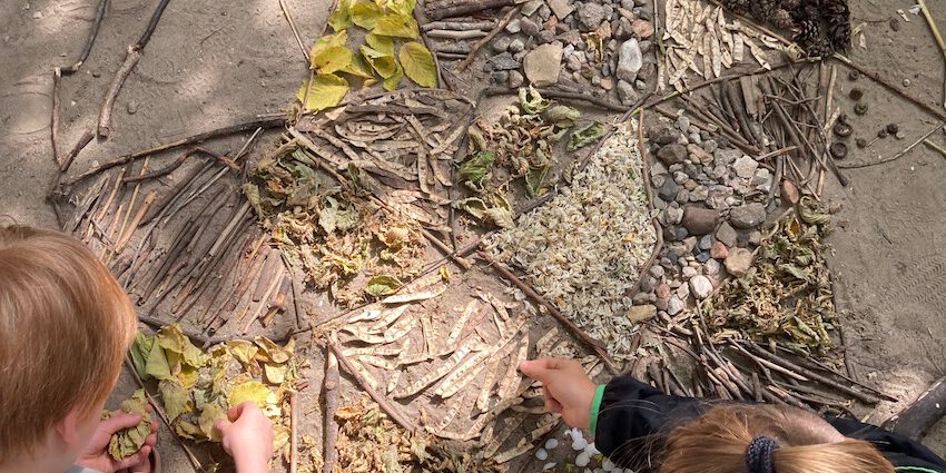 Projekt_LandArt_2023_2 Das Bild zeigt einen Blick auf sandigen Boden. Von oben sind zwei Kinder zu sehen, die ein Muster aus Pflanzenteilen, Stöcken und Blättern legen.
