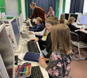 Das Bild zeigt mehrere Schülerinnen und Schüler im Computerraum einer Schule beim Arbeiten am Rechner. Im Hintergrund unterstützt eine Erwachsene einen Schüler.