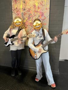 Zwei Kinder posieren mit E-Gitarren vor einem bunten Gemälde. Es handelt sich vermutlich um Mädchen, ihre Gesichter sind mit Emojis verdeckt.