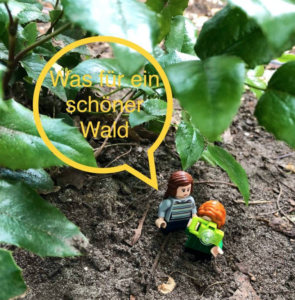 Unter einem Busch mit Blättern stehen zwei kleine Legofiguren auf dem Erdboden. Eine sagt zur anderen: Was für ein schöner Wald! 